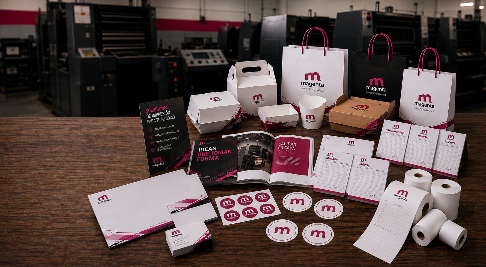 Trabajos impresos por Imprenta Magenta: bolsas de papel, cajas, folletos, talonarios y etiquetas sobre una mesa, con las máquinas de impresión al fondo.